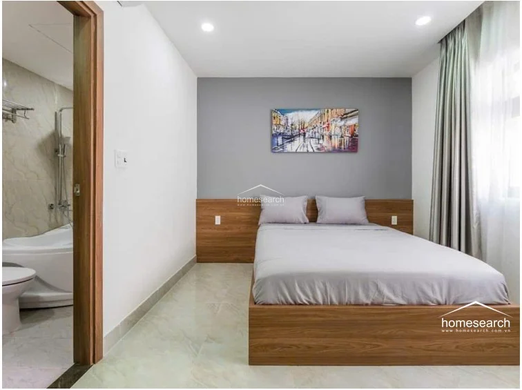 This bedroom features a platform bed with a matching wood headboard and a grey accent wall, creating a warm and inviting atmosphere. It includes an en-suite bathroom with marble-look wall tiles and a soaking tub, while the room itself is brightened by large windows draped in elegant floor-length curtains.