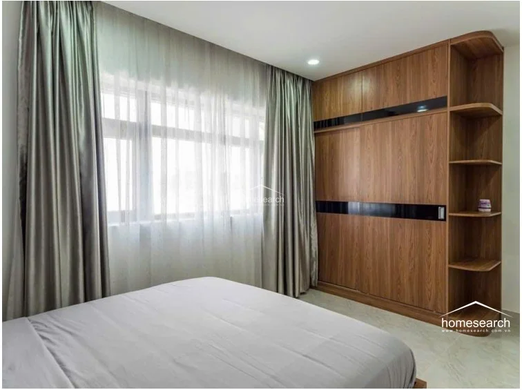 This bedroom features a large, floor-to-ceiling wooden wardrobe with integrated open shelving, providing ample storage. The space is brightened by a large window dressed in elegant, layered curtains, and the room is finished with light-colored tile flooring and a neutral color palette.