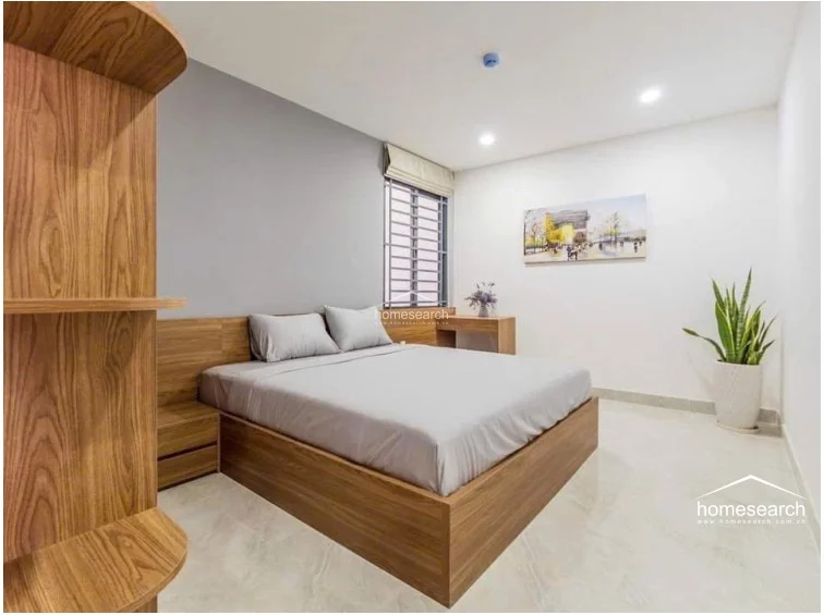 This bedroom layout centers on a wooden bed frame with a matching headboard and side table, set against a clean white and grey accent wall. The room includes a window with a Roman shade, light tile flooring, and a decorative plant, creating a minimalist and well-maintained atmosphere.