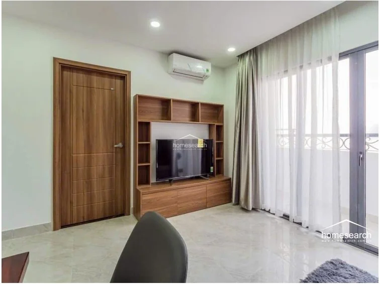 This living or bedroom area features a custom wooden media console with built-in shelving, providing functional storage for electronics and decor. A large sliding glass door leads to an outdoor balcony, allowing for significant natural light, while the room maintains a cohesive look with light tile flooring and wood-toned finishes.