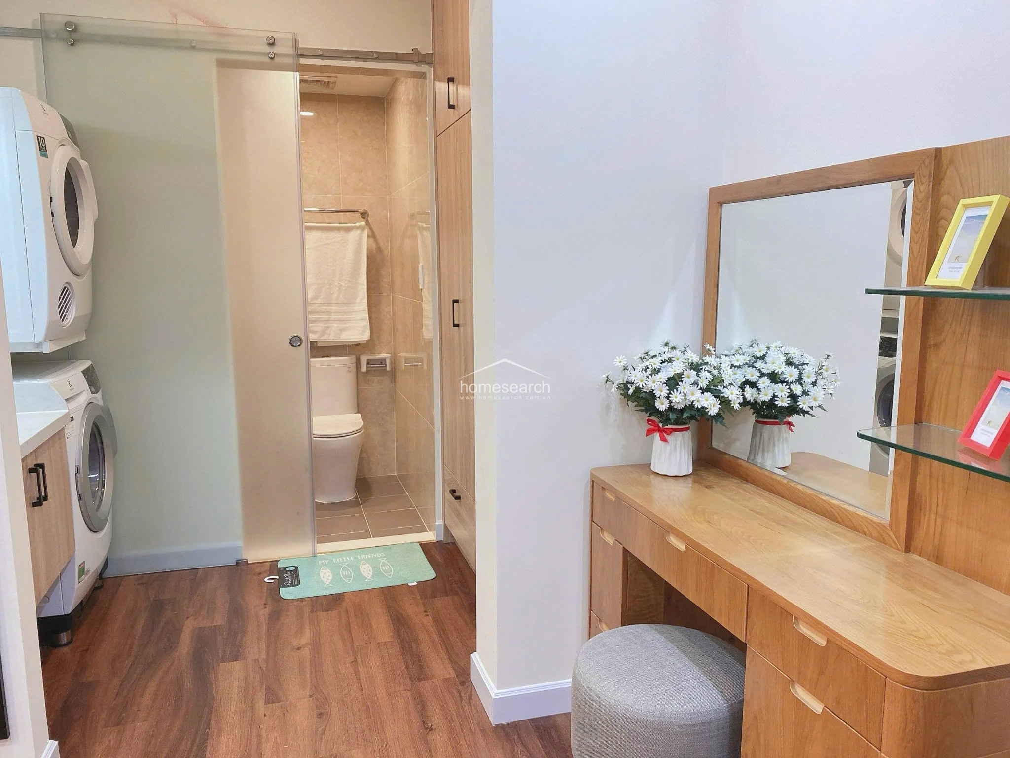 This functional dressing and laundry area connects seamlessly to the bathroom, featuring a stylish wooden vanity with a mirror and integrated shelving. A stackable washer and dryer unit is conveniently tucked into the space, maximizing efficiency in this compact, well-maintained layout.