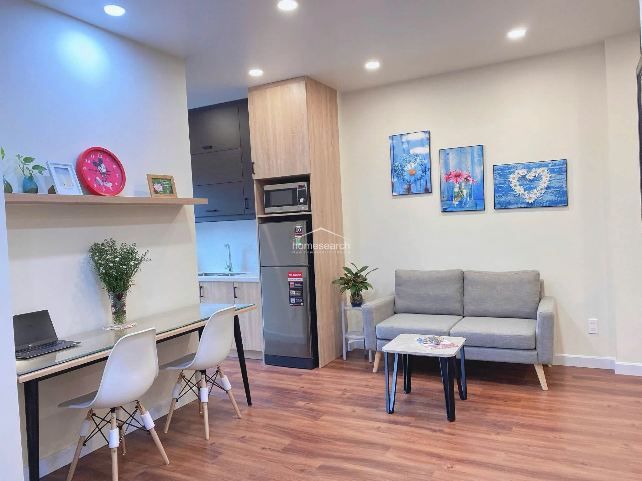 The living and work zone is efficiently furnished with a grey sofa, a small coffee table, and a glass-topped desk paired with two white modern chairs. A floating shelf above the desk provides decorative storage, while the overall aesthetic remains clean, well-maintained, and contemporary.
