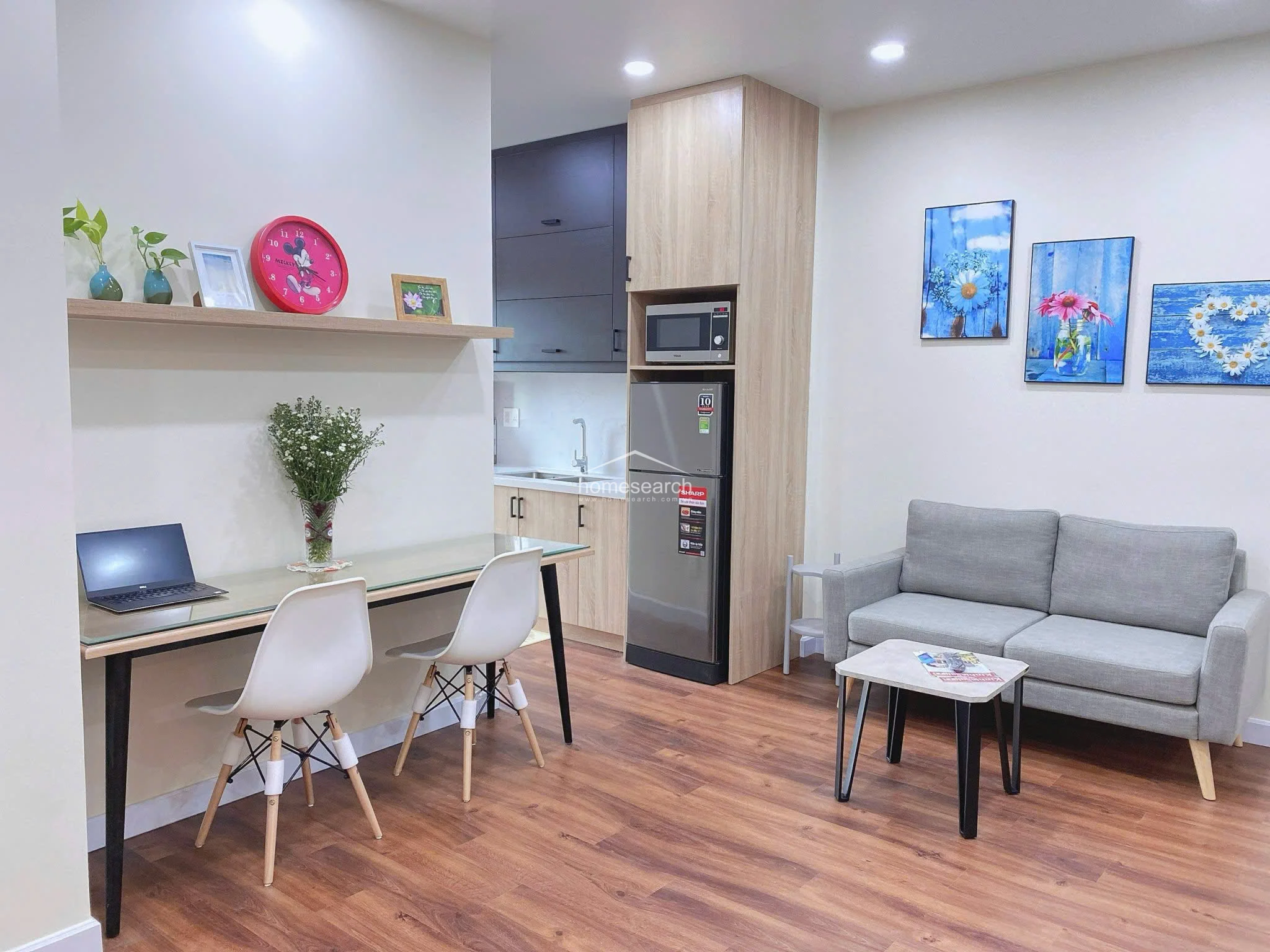 This view highlights the studio's efficient use of space, showing the transition from the sleeping area to the kitchen and balcony. The natural flow is enhanced by consistent flooring throughout and a bright, airy atmosphere provided by the balcony door.
