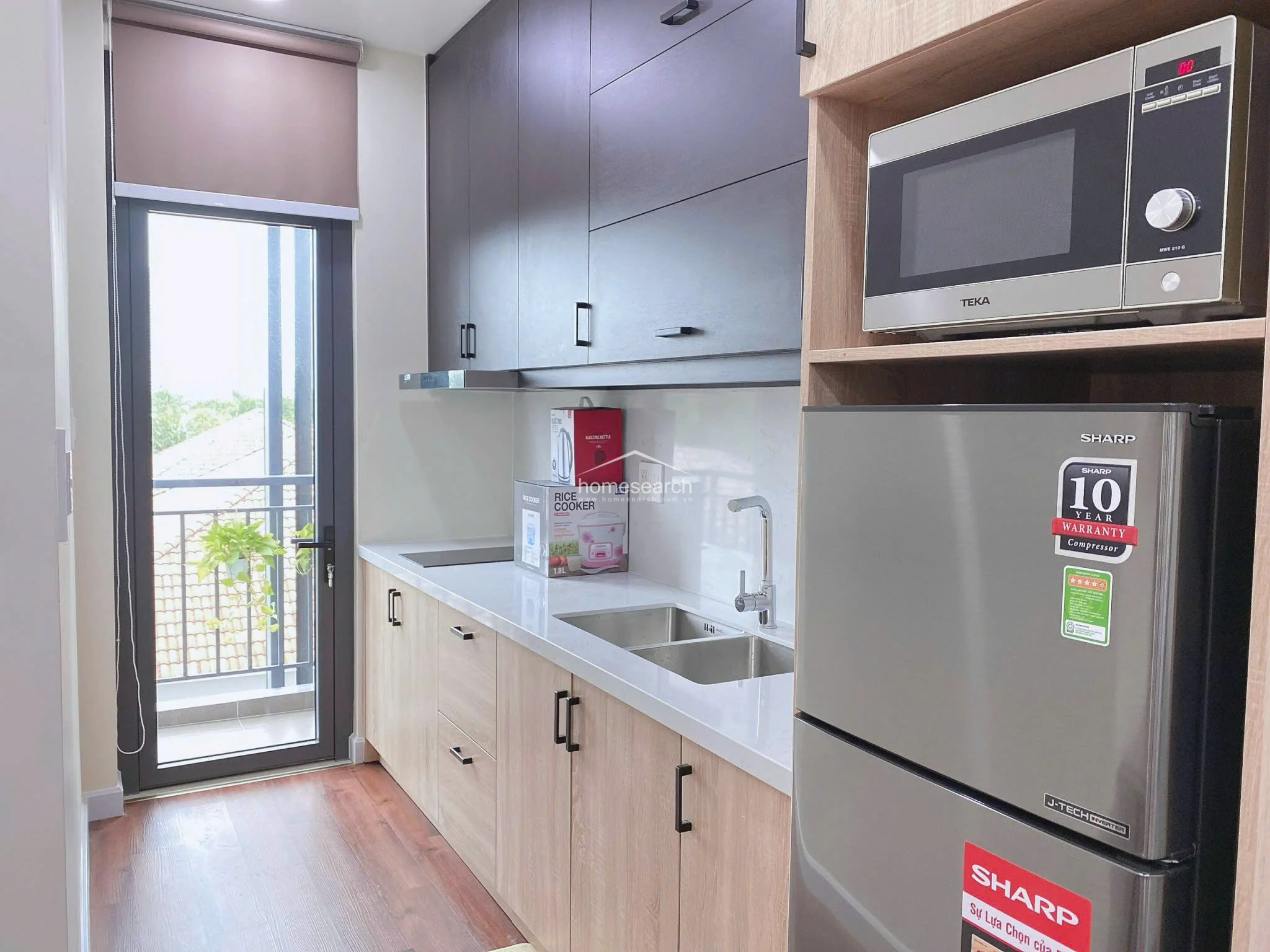 The compact kitchen is well-equipped with light wood lower cabinets, dark upper cabinetry, and a sleek white countertop. It includes a built-in Teka microwave, a Sharp refrigerator, and direct access to a balcony, which provides natural light and ventilation to the space.