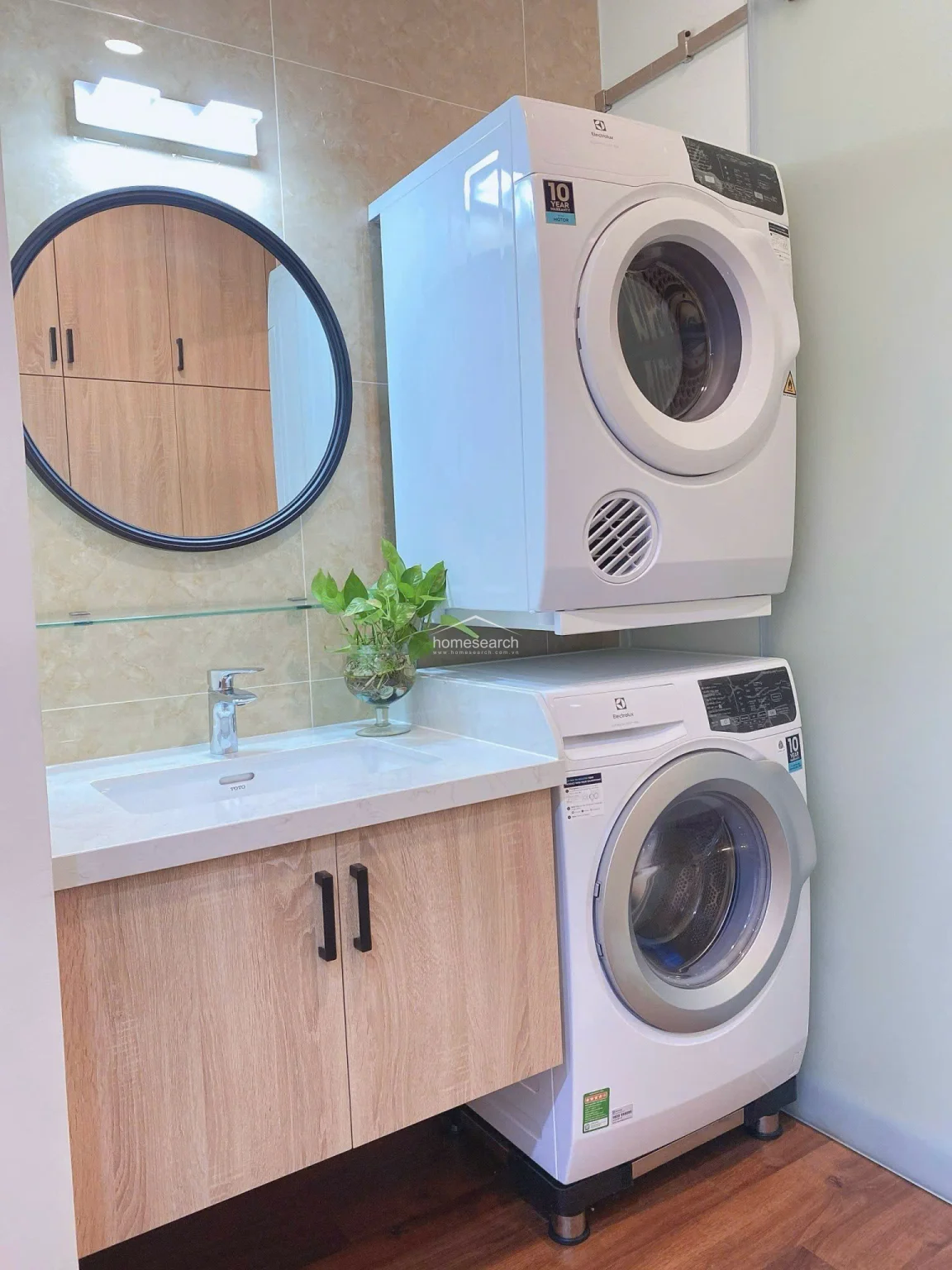 The bathroom vanity area is elegantly designed with a modern circular mirror, a quartz countertop, and a TOTO sink set against beige marble-look wall tiles. The space is highly functional, incorporating a built-in wooden cabinet underneath and a stacked laundry appliance setup for optimal space utilization.