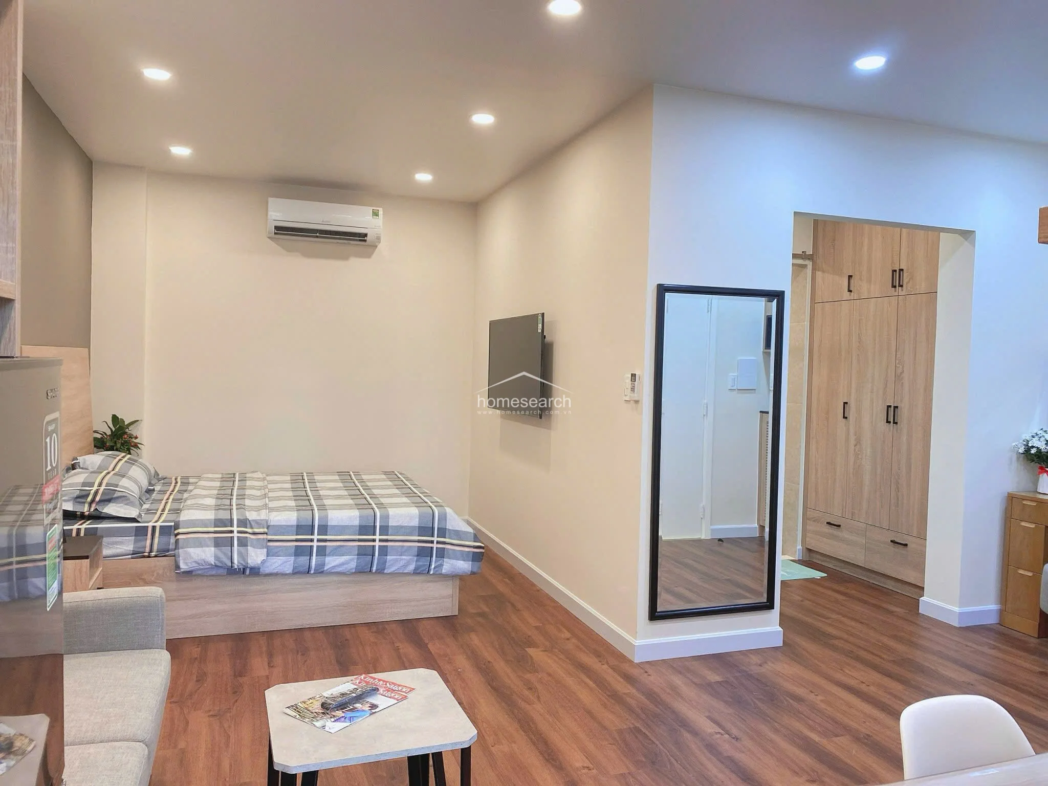 This studio bedroom features a clean, modern layout with warm wood-look flooring and neutral wall tones, creating a cozy and cohesive living space. The room is well-equipped with a wall-mounted AC unit, a flat-screen TV, and a built-in wooden wardrobe, while recessed ceiling lighting provides a bright and airy atmosphere.