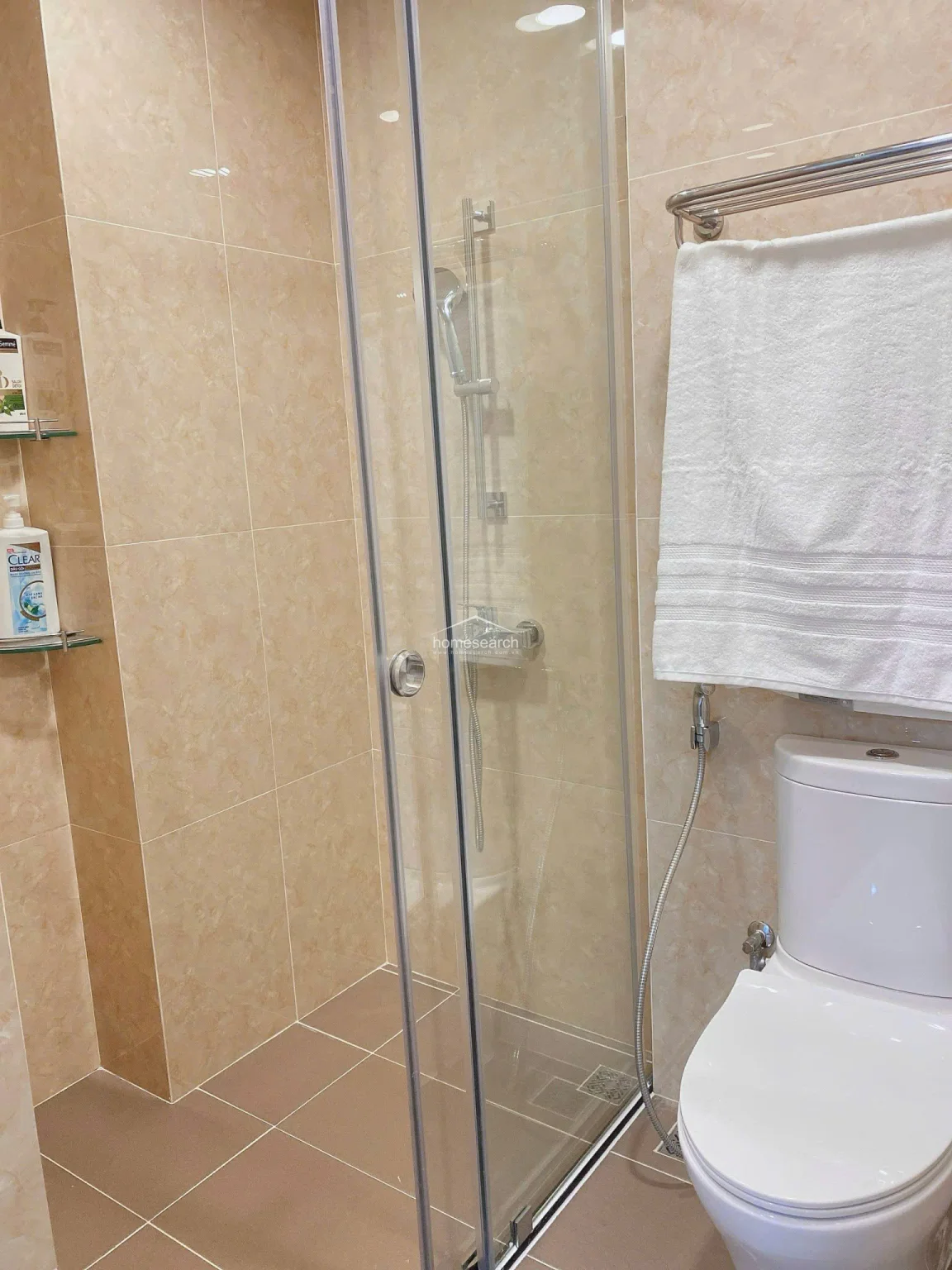 This modern bathroom features a sleek glass-enclosed shower stall and high-quality beige floor-to-ceiling tiling for a clean, upscale appearance. The space is well-maintained and includes essential fixtures like a wall-mounted towel rack, a handheld bidet sprayer, and a contemporary toilet, ensuring both comfort and convenience.