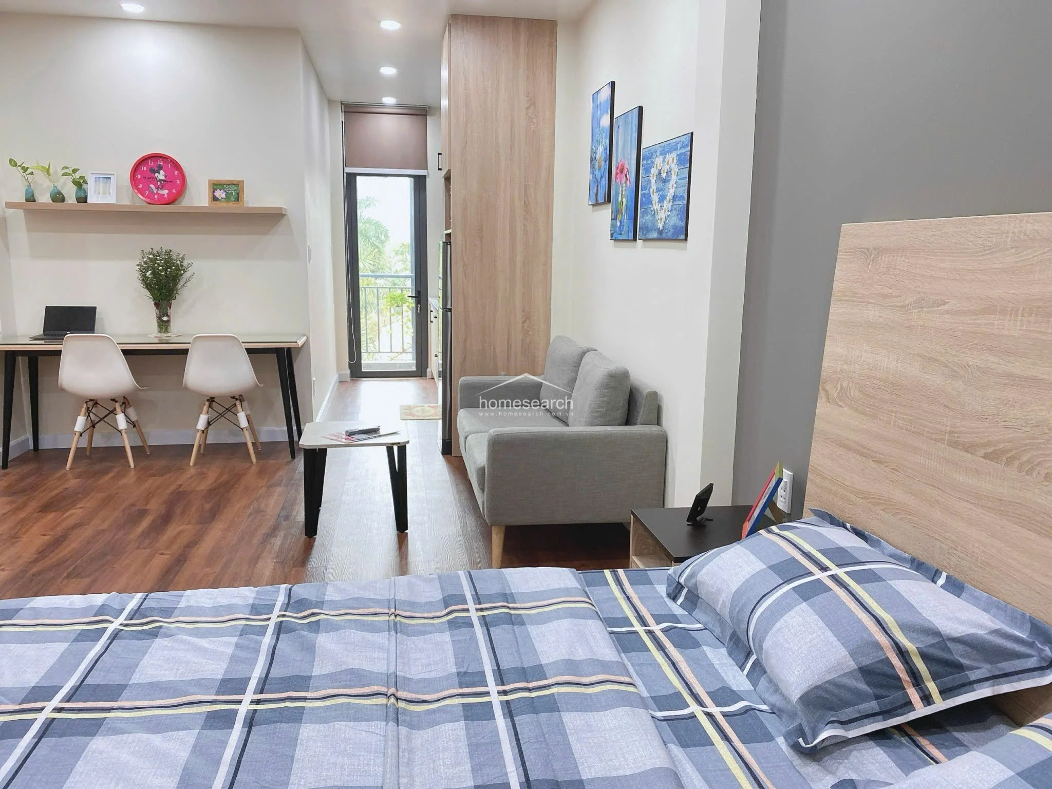This studio apartment features a functional open-plan layout that seamlessly integrates a sleeping area, a small lounge, and a workspace. The room is finished with warm wood-look flooring and neutral walls, accented by a grey feature wall behind the bed and modern recessed ceiling lighting.