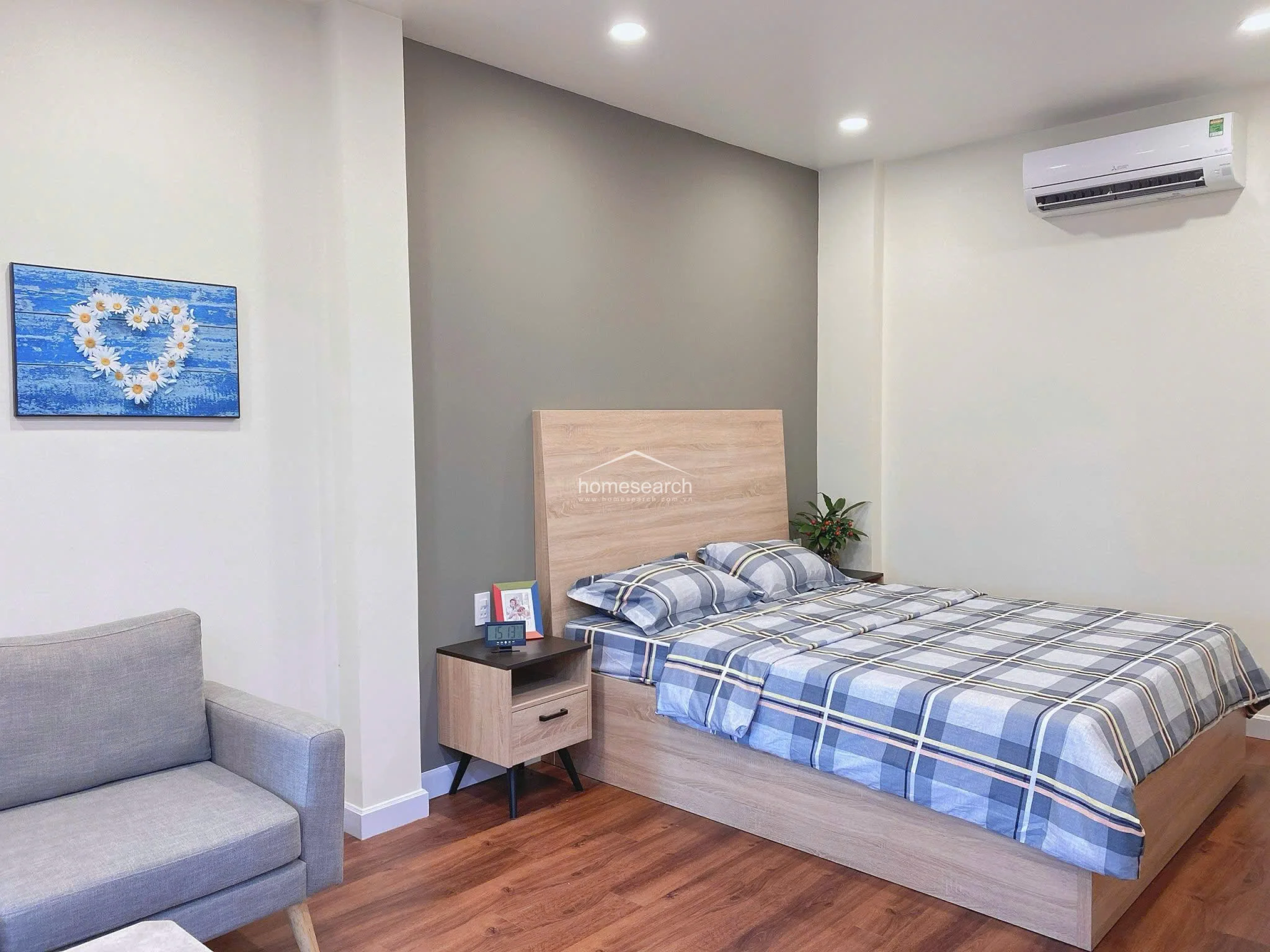 The sleeping area is anchored by a wooden bed frame with a matching headboard and side table, set against a sophisticated grey feature wall. The room is climate-controlled with a wall-mounted AC unit and maintains a cohesive, move-in-ready condition.