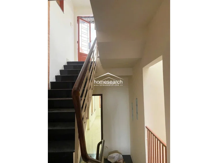 This staircase area features dark stone steps with a sturdy wooden handrail and metal balustrades, providing a clear vertical connection between the floors. The space is well-lit by natural light from a top-floor doorway and features minimalist wall finishes, maintaining a functional and clean aesthetic.