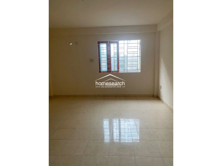 This bedroom is finished with simple, light-toned walls and matching tile flooring, maintaining a consistent design throughout the property. A window with metal security bars allows for natural light, and the room appears to be in good, well-maintained condition.