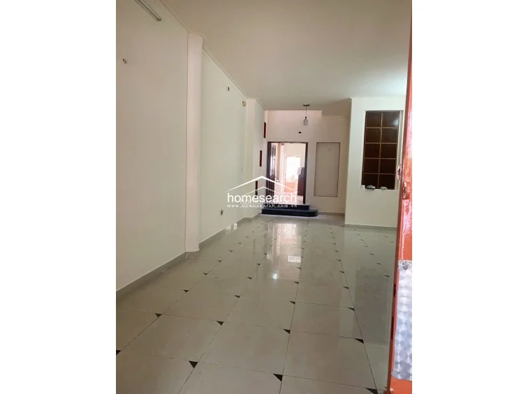 This spacious ground-floor living area features light-colored ceramic tile flooring and neutral white walls, creating a clean and open flow toward the rear of the property. The space is illuminated by a combination of natural light from the entrance and interior lighting, and it includes a dedicated recessed area for storage or display.