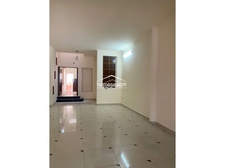 This spacious room features an open layout with light-colored tile flooring and a prominent vertical column. The space is illuminated by a wall-mounted light fixture and connects to an adjacent room through a doorway with a small step-up threshold.
