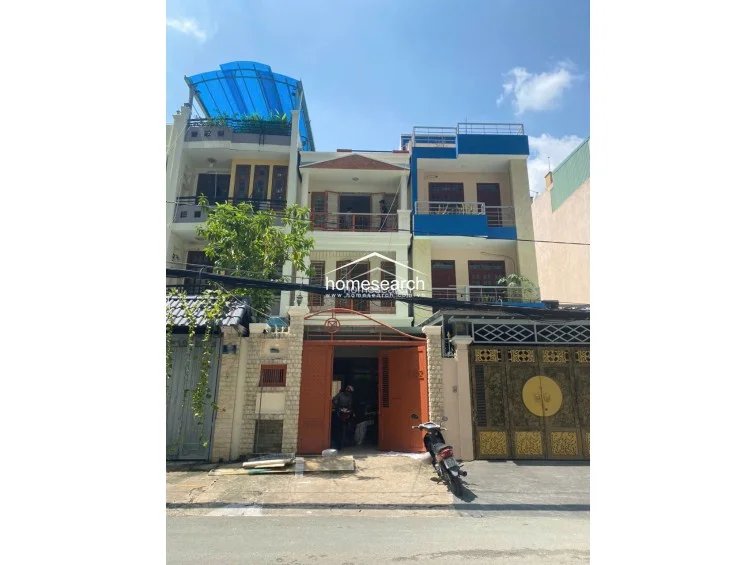 This three-story townhouse features a traditional facade with a gated ground-floor entrance, a second-floor balcony, and a rooftop terrace covered by a blue canopy. The exterior is finished in a mix of light-colored plaster and decorative brickwork, showing signs of standard residential wear and tear.
