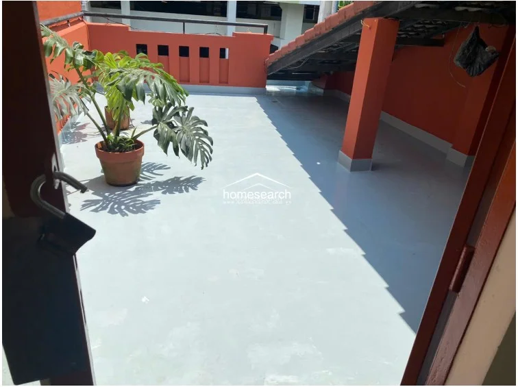 This outdoor terrace offers a private, open-air space finished with light grey flooring and accented by vibrant orange walls. It includes a sheltered area under a pitched roof and is currently decorated with a large potted Monstera plant, providing a clean and well-maintained outdoor retreat.