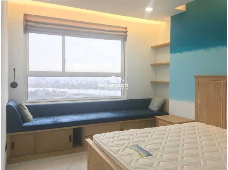 This view highlights the bedroom's functional storage solutions, including a long, upholstered blue window seat with built-in wooden cabinets below. The space is efficiently organized with light-wood floating shelves and a simple, modern color palette that emphasizes the expansive urban view.