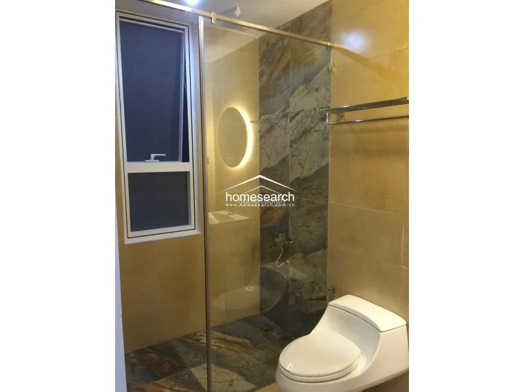 This modern bathroom features a sleek glass-enclosed shower stall with striking, large-format marble-patterned wall tiles that contrast against the warm, solid-colored beige tiles. The space is equipped with a contemporary white toilet, a wall-mounted towel rack, and a backlit circular mirror that adds a sophisticated touch to the well-maintained interior.