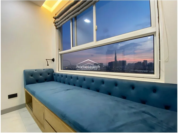This bedroom features a sophisticated window-side lounge area with a custom blue tufted velvet bench seat that includes built-in wooden storage drawers underneath. The large window provides an expansive, unobstructed city skyline view, complemented by modern Roman shades and warm recessed ceiling lighting.