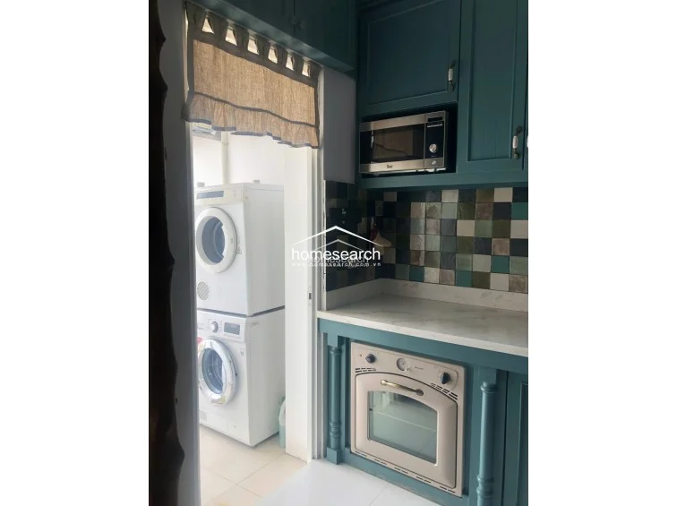 The kitchen showcases a vibrant teal cabinetry scheme paired with a multi-colored mosaic tile backsplash and white stone countertops. It is equipped with a built-in oven and microwave, and it connects directly to a utility area housing a stacked washer and dryer unit.