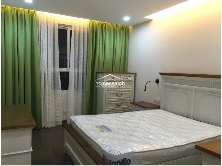 This bedroom is decorated in a classic style with a white wooden bed frame, matching nightstands, and a tall chest of drawers. The space is softened by floor-to-ceiling green and white patterned curtains, while the overall condition appears well-maintained and tidy.