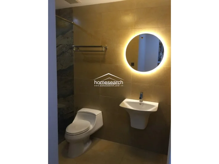 This bathroom features warm-toned wall tiles, a modern wall-mounted sink, and a circular LED-backlit mirror that serves as a focal point. The space is well-maintained and includes a glass-enclosed shower area with darker accent tiling for a contemporary aesthetic.