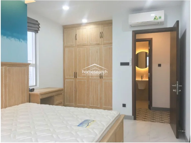This bedroom features a clean, modern layout with light-colored tile flooring and a striking blue ombre accent wall. It includes a custom light-wood wardrobe, a matching bed frame, and a wall-mounted AC unit, with an open doorway leading to a bathroom that features a backlit circular mirror.