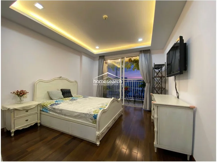 This master bedroom showcases a sophisticated aesthetic with rich dark wood flooring and a classic, ornate bed frame. The room flows directly onto the balcony through large sliding glass doors, and the ceiling is finished with elegant recessed lighting and a cove feature.