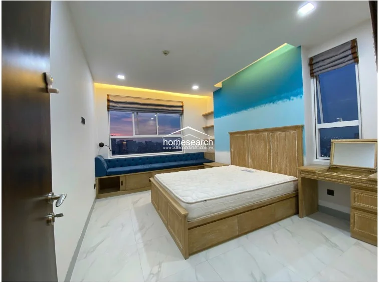 This bedroom is designed for comfort, centered around a large bed and a unique built-in window bench with integrated storage underneath. The room is brightened by natural light from the window, complemented by a decorative blue accent wall, wooden floating shelves, and a dedicated vanity area.