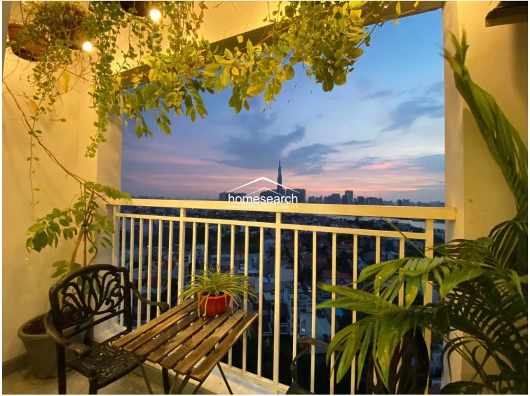 This balcony offers a serene outdoor retreat, featuring a small bistro set and lush potted greenery that frames a stunning sunset view of the city skyline. The space is well-maintained and provides a perfect, intimate connection to the outdoors, enhanced by warm overhead lighting.