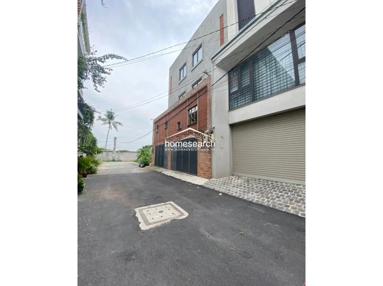 This exterior view shows a multi-story modern property with a paved driveway and a secure, gated entrance. The architecture features a blend of smooth grey concrete and decorative brickwork, with large windows providing ample natural light to the upper levels.