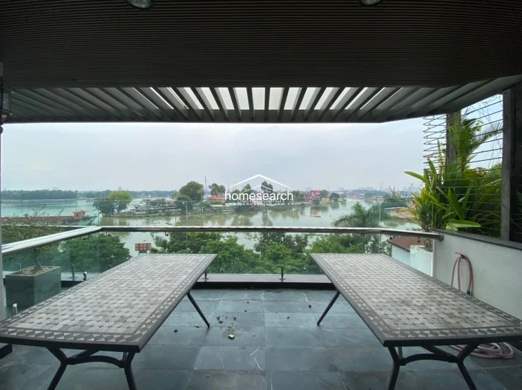This expansive outdoor terrace features a covered area with a slatted roof, providing partial shade over two large mosaic-tiled tables. The space offers a stunning, unobstructed panoramic view of the river and surrounding landscape, framed by a modern glass railing.