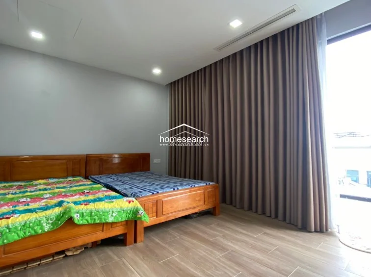 This bedroom features a functional layout with two wooden beds and light-colored walls, creating a simple and clean aesthetic. The room is equipped with recessed ceiling lighting and a large window covered by floor-to-ceiling brown curtains, which provide privacy and effective light control. The flooring consists of light wood-tone tiles, and the overall condition appears to be well-maintained and move-in ready.