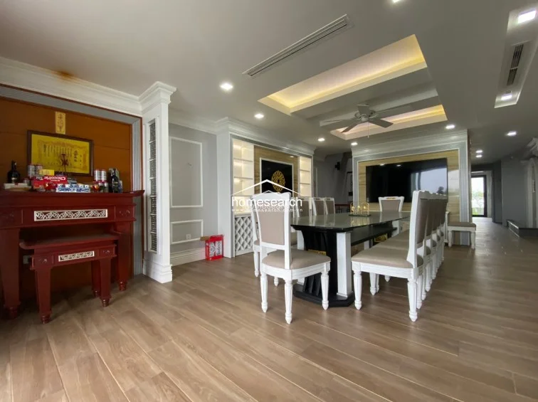 This spacious dining and living area features elegant wood-look flooring and a sophisticated tray ceiling with integrated lighting and a ceiling fan. The room is thoughtfully laid out with a formal dining set, a built-in wine rack, and a traditional altar, creating a balanced flow between functional living space and cultural elements.