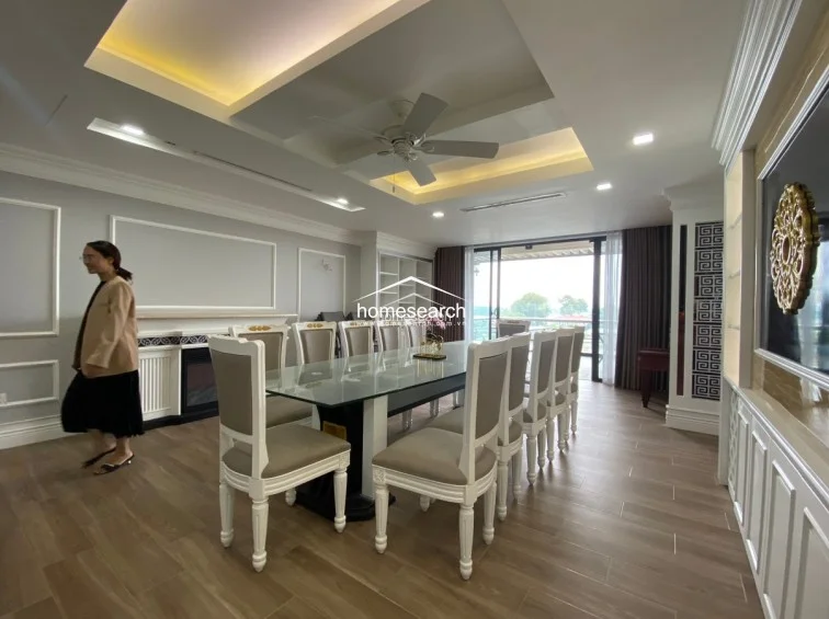 This elegant dining area features a long glass-topped table with high-backed, cream-upholstered chairs, perfect for formal gatherings. The space is finished with wood-look tile flooring, recessed ceiling lighting with a central fan, and a neutral color palette that flows seamlessly toward a balcony with an outdoor view.