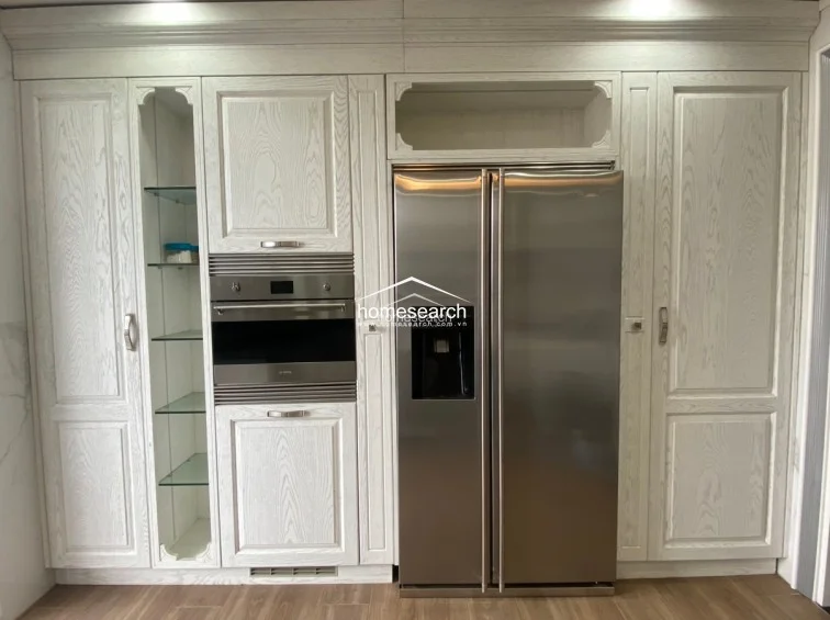 This high-end kitchen wall unit showcases custom white-washed wood cabinetry with integrated glass shelving and a professional-grade stainless steel double-door refrigerator. The design emphasizes clean lines and premium storage, featuring a built-in oven and a seamless, organized aesthetic that maintains the property's luxurious tone.
