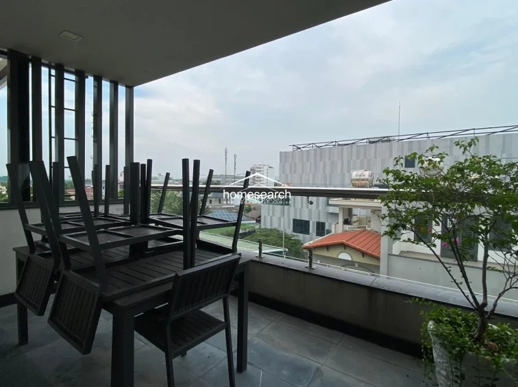 This covered balcony serves as an outdoor dining or lounge area, featuring dark-toned patio furniture and a view of the surrounding urban landscape. The space is finished with slate-style tiling and glass railings, providing a seamless transition from the interior to the outdoors.
