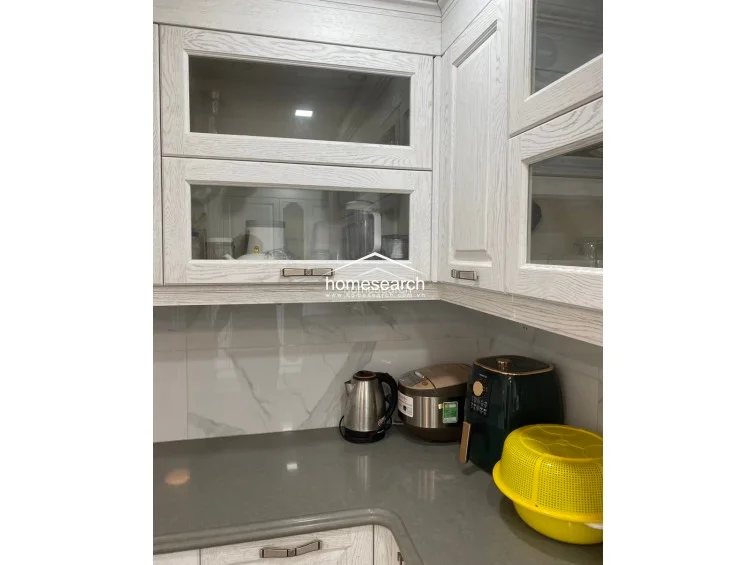 This kitchen section showcases white, wood-grain cabinets with glass inserts and a durable grey stone countertop. The space is well-equipped with modern small appliances, including a kettle and air fryer, set against a clean, white marble-patterned backsplash.