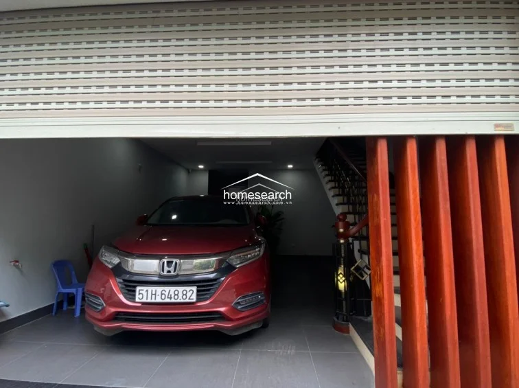 This ground-floor garage area serves as a secure parking space, accessed by a modern, motorized roll-up metal shutter. It features clean, neutral walls and tiled flooring, with an open-riser wooden staircase leading to the upper levels of the home.
