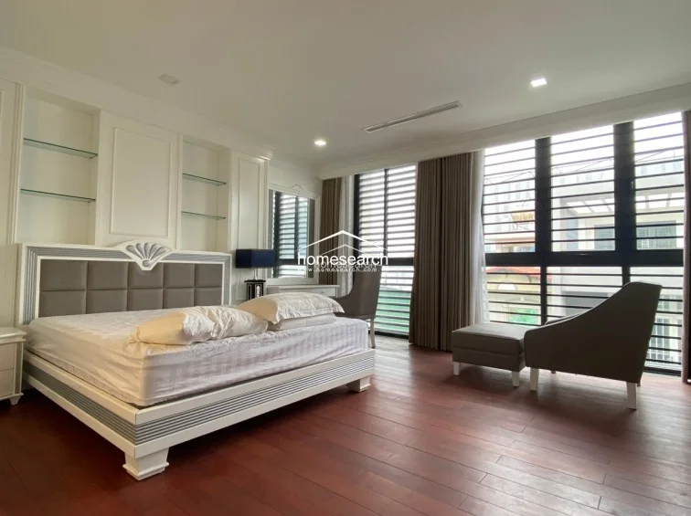 This spacious bedroom is elegantly designed with dark wood flooring and a large window featuring horizontal blinds and brown curtains for ample natural light. It includes a modern bed with a grey upholstered headboard, built-in white shelving, and a dedicated vanity area, offering a sophisticated and organized living space. The room is in excellent condition, showcasing high-quality finishes and a cohesive, neutral color scheme.