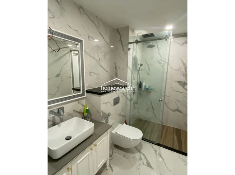 This modern bathroom is finished with high-end white marble-patterned tiles on both the walls and floor, creating a luxurious and bright appearance. It is equipped with a contemporary vessel sink, a wall-mounted toilet, and a glass-enclosed shower area with a rainfall showerhead. The fixtures are in excellent condition, and the overall design reflects a high level of maintenance and modern style.