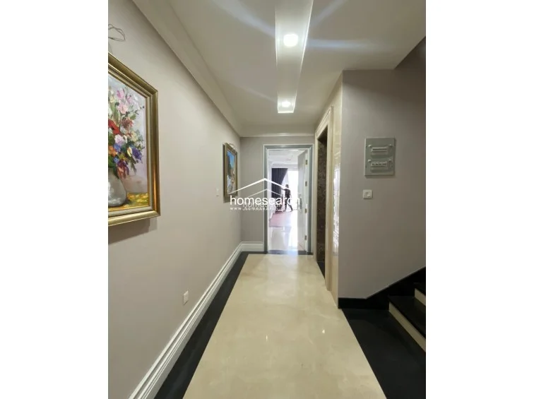This hallway serves as a clean, transitional space featuring polished light-colored marble flooring with a dark border, leading directly into the bedroom area. The walls are painted in a soft neutral tone and decorated with framed artwork, while the ceiling includes recessed lighting for a bright, welcoming feel. The architectural details, such as the crown molding and clean lines, indicate a well-maintained and upscale property.