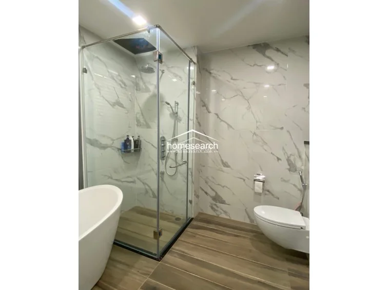 This modern bathroom features high-end marble-effect wall tiling and contrasting dark wood-look floor tiles. It is equipped with a contemporary freestanding soaking tub, a glass-enclosed walk-in shower with rainfall fixtures, and a sleek wall-mounted toilet.