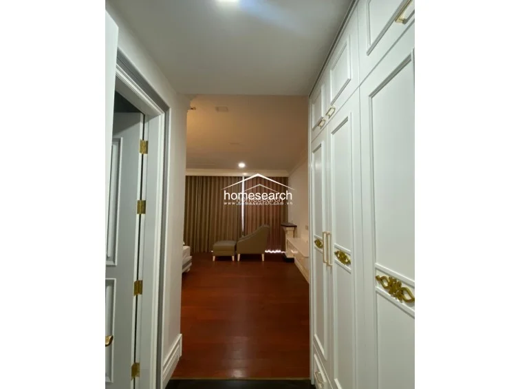 This image shows a transitional hallway area leading into a bedroom, featuring floor-to-ceiling white built-in wardrobes with elegant gold hardware. The flooring consists of rich, dark hardwood, creating a warm and sophisticated aesthetic that flows seamlessly into the main living space.