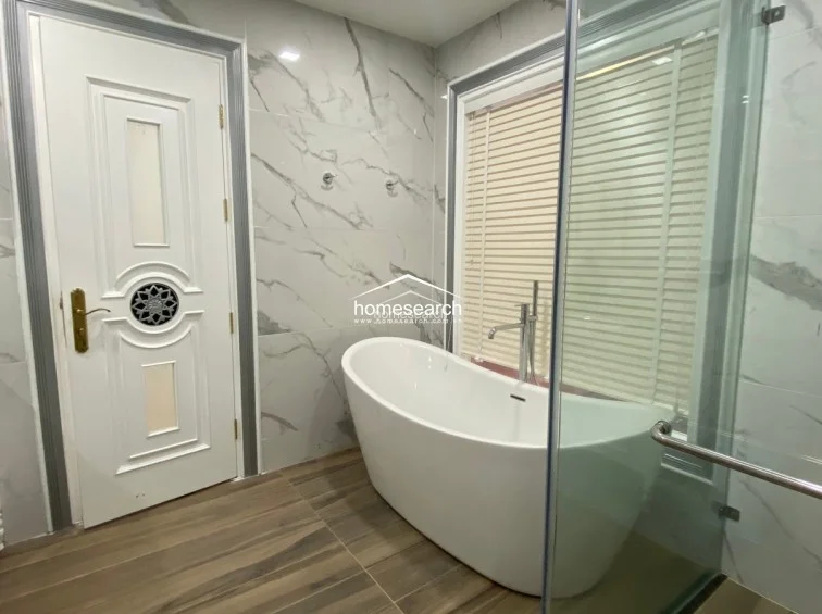 This luxurious bathroom features a contemporary freestanding soaking tub positioned next to a window with adjustable blinds for privacy. The room is finished with high-end white marble-vein wall tiles and warm wood-look flooring, creating a sophisticated and spa-like atmosphere.