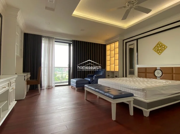 This spacious bedroom is elegantly designed with dark hardwood flooring, a coffered ceiling with recessed lighting, and a ceiling fan. The room is furnished with a plush bed, a blue leather bench, and a matching armchair, all complemented by floor-to-ceiling curtains and custom built-in shelving.