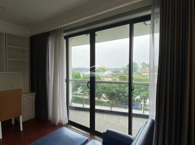 This view from the bedroom highlights the seamless transition to the outdoor balcony through large, full-height sliding glass doors. The space is framed by heavy, high-quality drapes and includes a dedicated study area with built-in shelving, emphasizing a functional and well-maintained layout.
