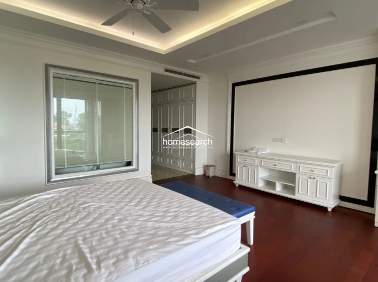 This spacious bedroom features rich dark wood flooring and a sophisticated tray ceiling with recessed lighting and a central ceiling fan. It includes a large white media console, a blue upholstered bench, and extensive built-in white cabinetry for ample storage.