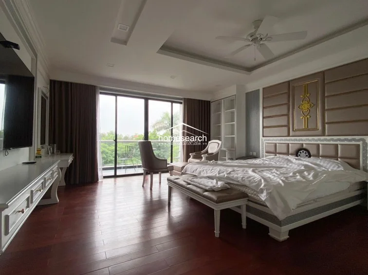 This master bedroom suite is elegantly furnished with a large bed featuring a tufted, neutral-toned headboard and gold accents, complemented by a matching vanity and chair. Large sliding glass doors provide access to a private balcony, while the room is finished with dark wood flooring and recessed ceiling lighting.