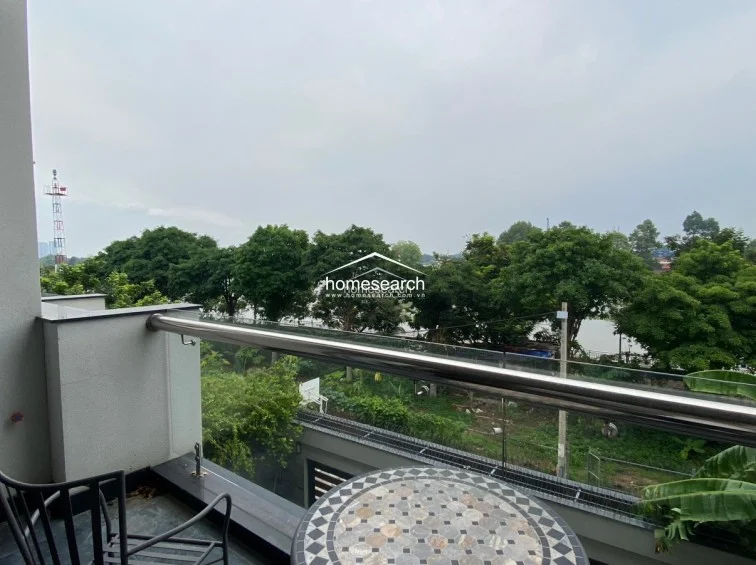 This private balcony offers a peaceful outdoor retreat with a small mosaic-topped bistro table and a view overlooking lush greenery and a nearby waterway. The space is enclosed by a modern glass and stainless steel railing, providing a clean, unobstructed look at the surrounding landscape.