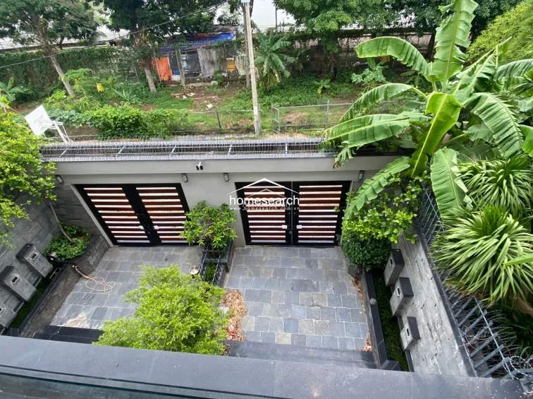 This elevated view captures a private, enclosed courtyard paved with slate-grey stone tiles, featuring lush greenery and banana trees that provide a tropical feel. The space is secured by two modern, horizontal-slat metal gates and high perimeter walls, offering a secluded and well-maintained outdoor area.